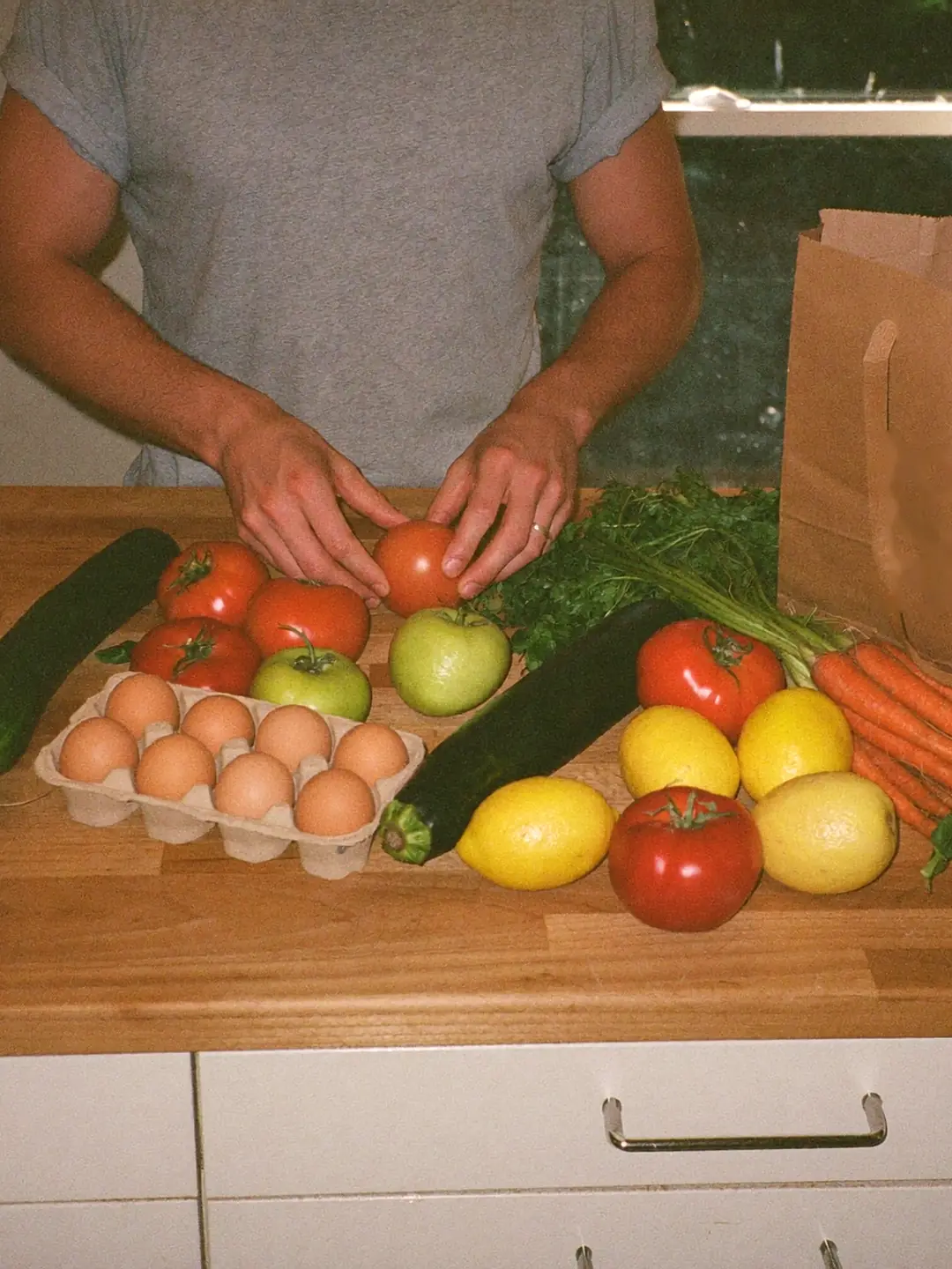 Des courses posées sur un plan de travail : tomates, courgettes, citrons, carottes, salade, œufs et un sac en papier kraft.