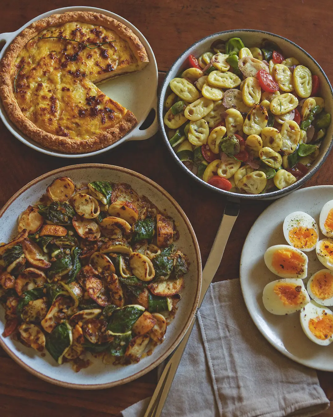 Plusieurs plats maison posés sur une table en bois : une quiche, des œufs mimosa, une salade de pâtes et un gratin.